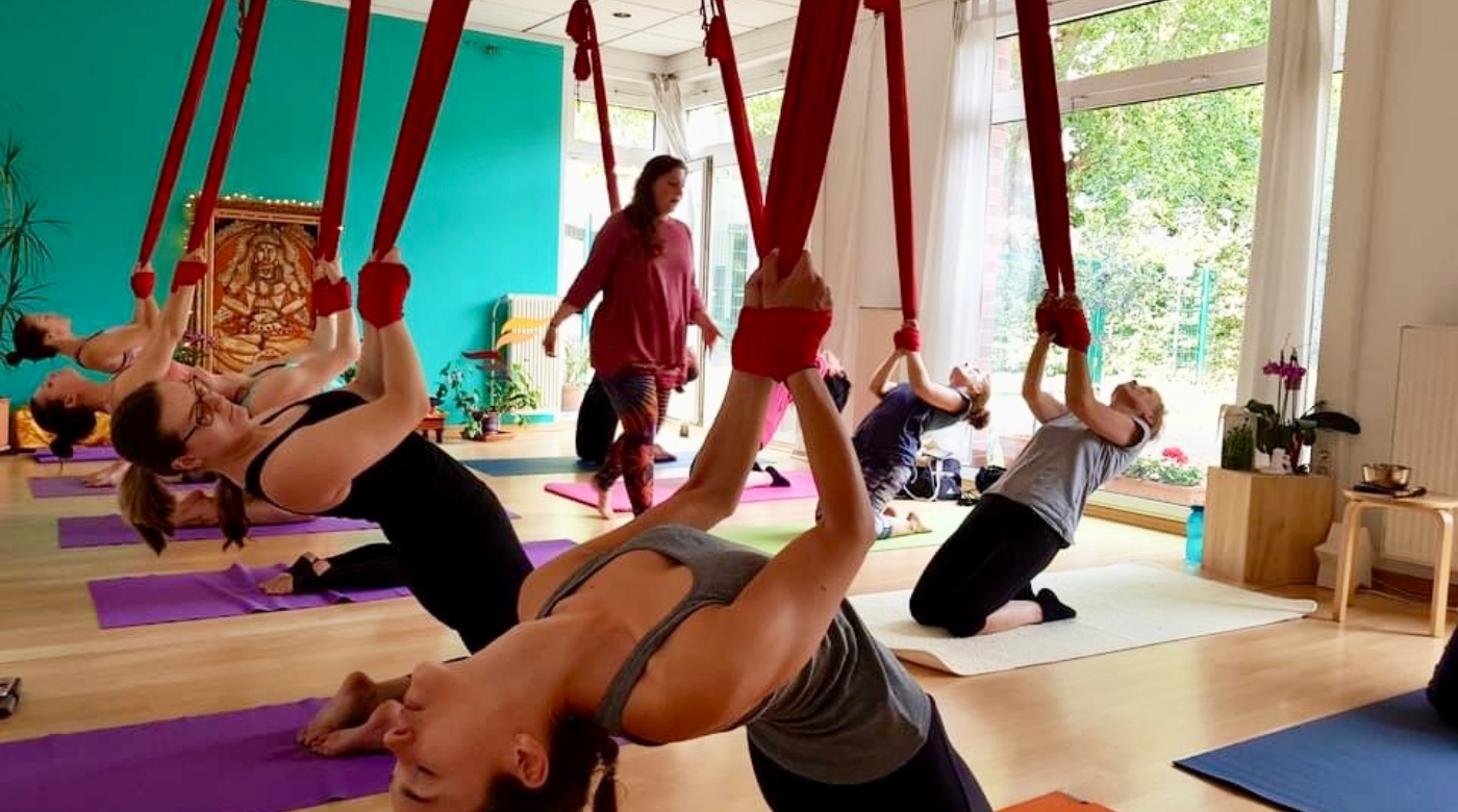 Teilnehmerinnen praktizieren Aerial Yoga mit roten Tüchern in einem hellen Yogaraum.