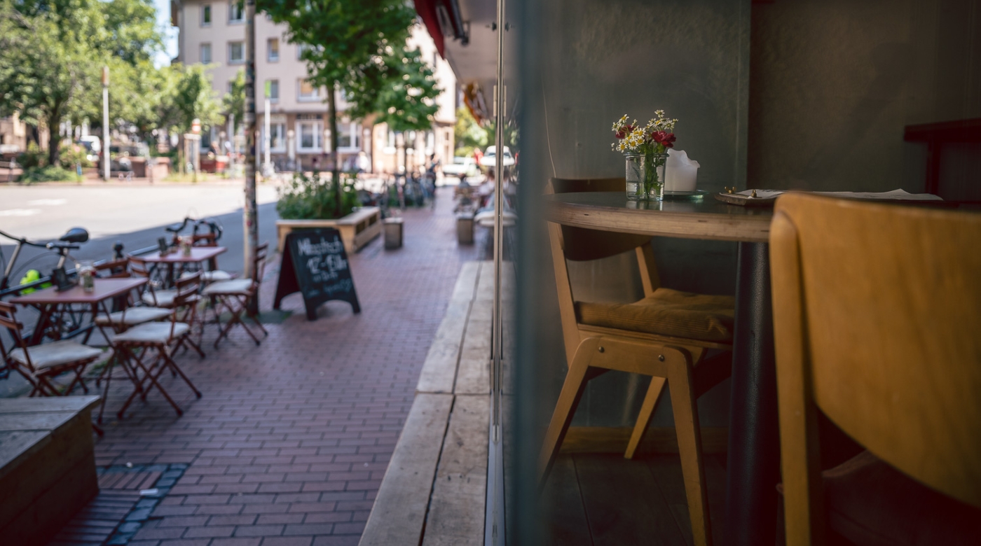 Café mit Innen- und Außenbereich, Holztisch mit Blumen, Ziegelpflaster und Straßenszene.