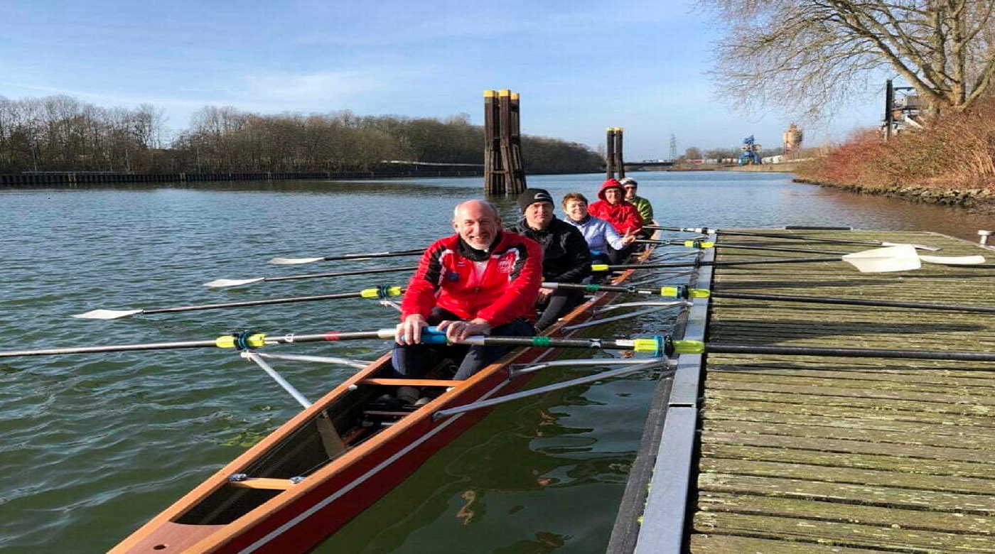 Ruderteam in einem Boot auf einem Fluss, angelegt an einem hölzernen Steg.