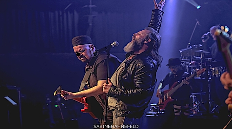 Drei Musiker auf einer Bühne mit Lichteffekten und Instrumenten im Hintergrund.