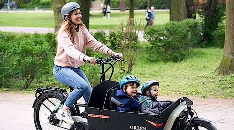 Lächelnde Frau mit Helm mit Lastenrad und 2 Kindern mit Helm